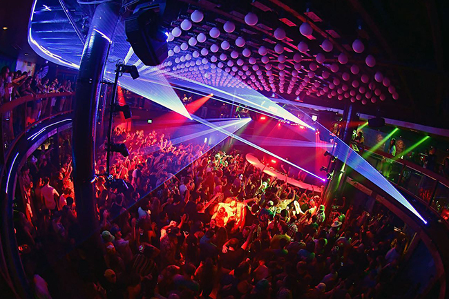 An elevated view of District’s dance floor with guests along a curved balcony railing at the left, ceiling spheres and green lasers